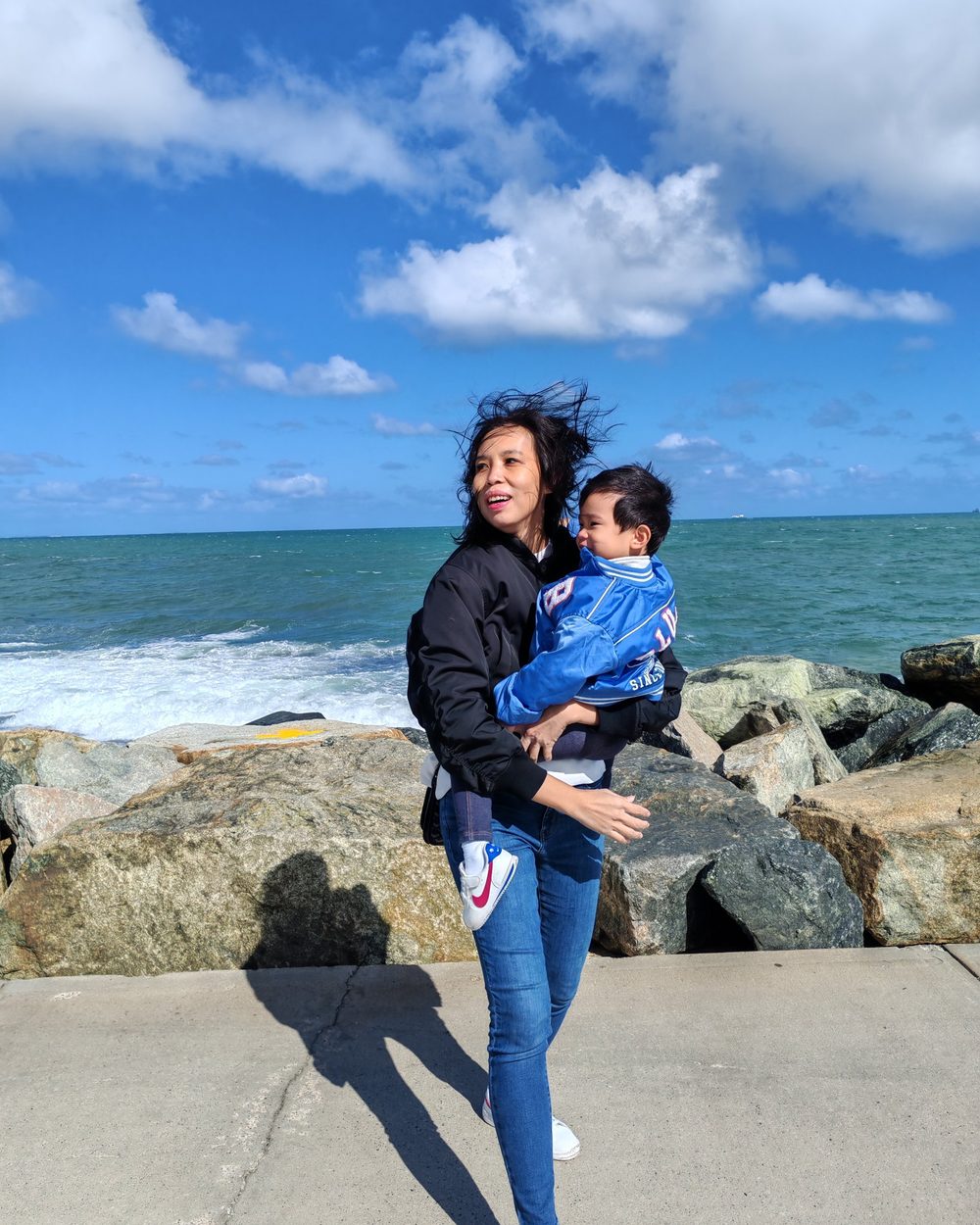 Joan carrying her little one by the sea, hair blowing in the wind